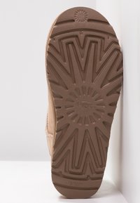 Semelle d'une botte UGG beige avec un motif de rayons de soleil et le logo "UGG" centré sur la semelle extérieure en caoutchouc.