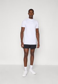 Weißes sportliches T-Shirt mit kurzen Ärmeln und rundem Ausschnitt, kombiniert mit schwarzen Shorts und weißen Sneakern, vor einem neutralen Hintergrund.