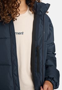 Persona con capelli ricci che indossa una camicia beige parzialmente coperta da una giacca trapuntata blu navy con zip aperta e una tasca verticale con zip.