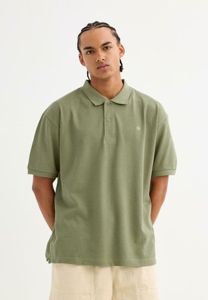 Camisa polo verde oliva hecha de tela suave, con un cuello clásico, dos botones, mangas cortas y un pequeño logo en el pecho.