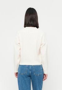 Pulover bej deschis cu un model texturat, guler rotund și manșete cu nervuri, combinat cu blugi albaștri cu talie înaltă.