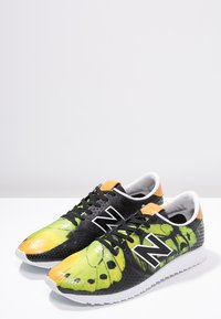 Baskets de sport vertes et noires avec un motif de ailes de papillon, semelle blanche, matériau texturé et lacets noirs. Accents orange au niveau du talon.
