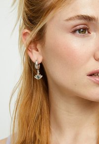 Créole en argent avec un pendentif en forme de couronne orné d'une pierre précieuse verte. Texture lisse et finition brillante. Portée à l'oreille d'un modèle.