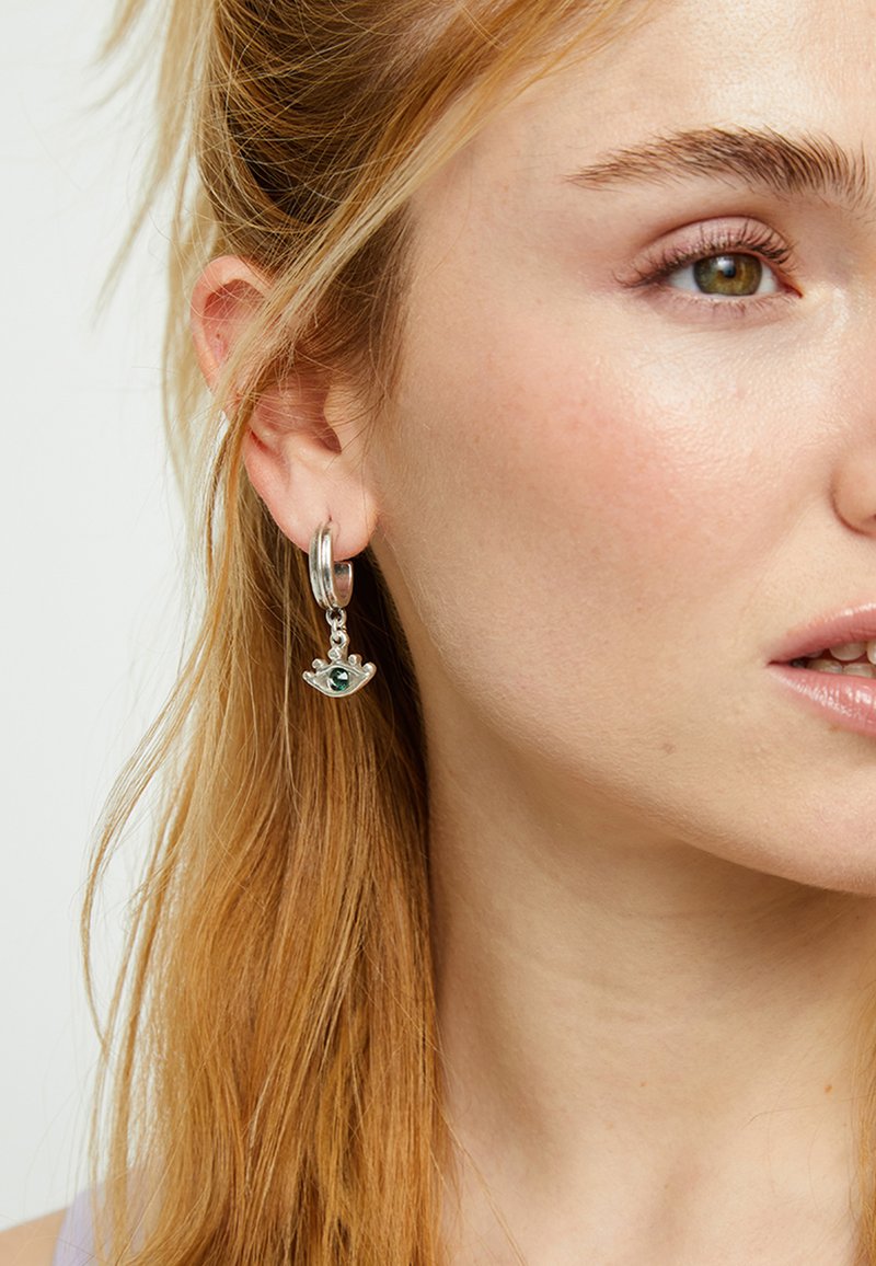 Créole en argent avec un pendentif en forme de couronne orné d'une pierre précieuse verte. Texture lisse et finition brillante. Portée à l'oreille d'un modèle.