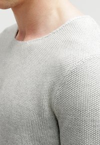 Detailní záběr na rameno a krk osoby, která má na sobě svetřík ze světlého šedého strukturovaného úpletu s kulatým výstřihem.