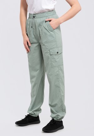 Osoba ubrana w jasnozielone spodnie typu cargo ze ściągaczami u kostek oraz czarne trampki, stojąca z jedną ręką w kieszeni i białą bluzką.