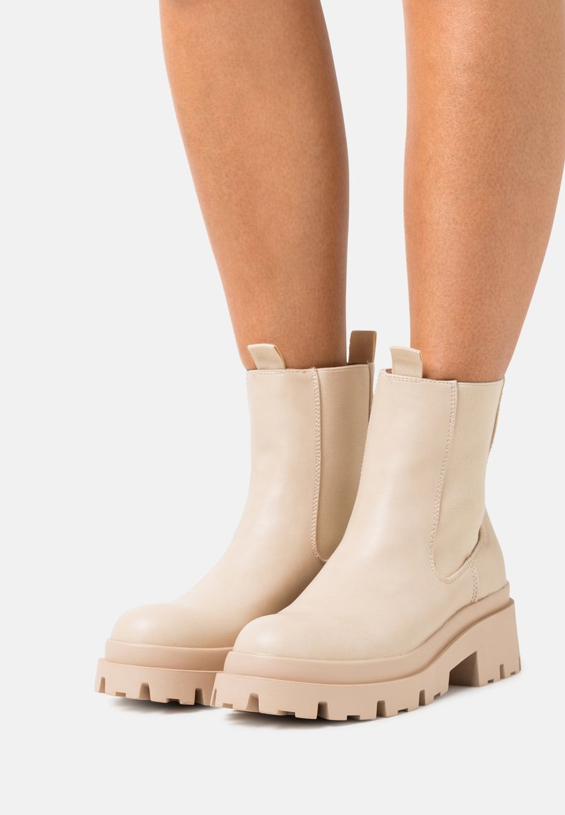 Bottes beiges jusqu'à la cheville avec semelles à crampons épaisses, portées sur des jambes bronzées, dotées de languettes de tirage et de panneaux élastiques latéraux pour un enfilage facile.
