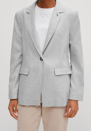 Personne portant un blazer gris clair ajusté sur une chemise blanche et un pantalon beige, debout avec les bras détendus le long du corps.