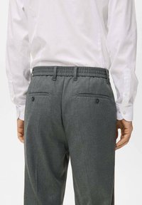 Pantalons ajustés gris avec un tissu texturé, une taille élastique et deux poches arrière avec des fermetures à boutons. Porté avec une chemise blanche.