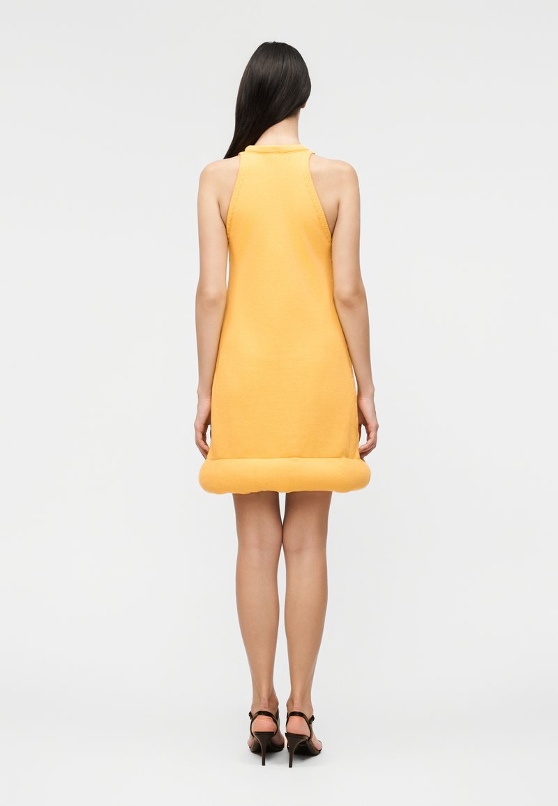 Woman with long dark hair wearing a sleeveless yellow dress with a rolled hem, standing back, paired with black high-heeled sandals.