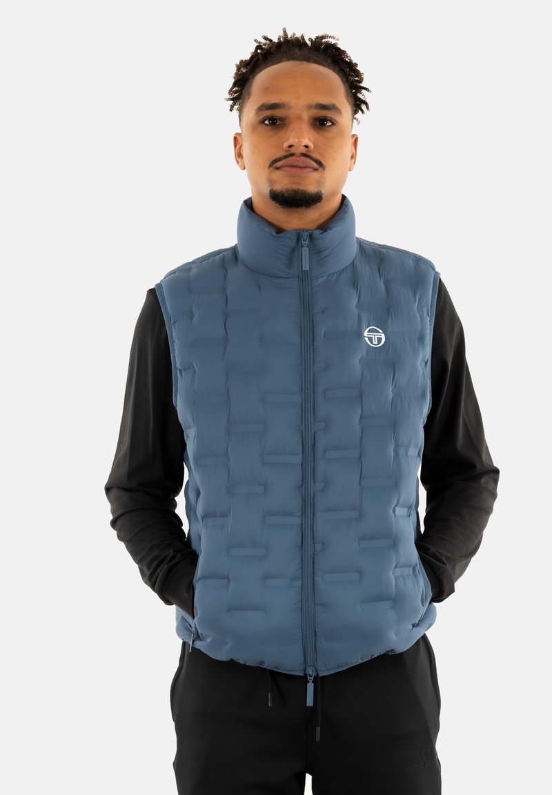 Gilet imbottito blu con colletto alto, chiusura con zip e superficie testurizzata. Indossato sopra una maglietta a maniche lunghe nera, con dettaglio logo a contrasto.