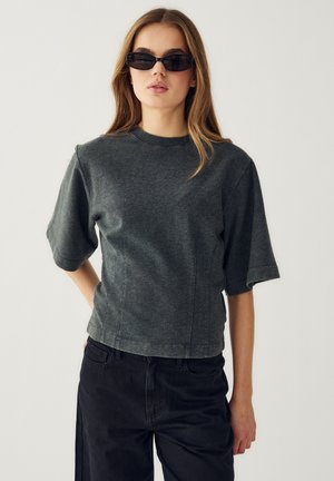 Femeie purtând un top cu mânecă scurtă gri închis, pantaloni negri și ochelari de soare negri înguști, stând în fața unui fundal simplu, deschis la culoare.