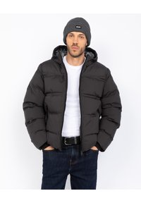 Sort polstret jakke med hætte, med lynlås og elastiske manchetter. Båret over en hvid t-shirt og kombineret med mørkeblå jeans.