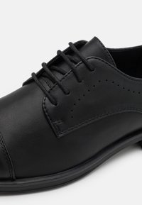 Chaussure habillée en cuir noir avec une texture lisse, dotée d'un laçage fermé, de détails perforés sur le côté et d'un bout arrondi.