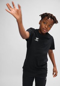 Camiseta de algodón negra con mangas cortas y cuello redondo, con un logo blanco en el lado izquierdo del pecho, combinada con pantalones negros.