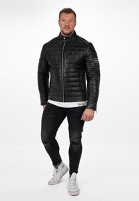 Zwarte gestikte leren jas met hoge kraag, voorkant met ritssluiting en patroonaccenten. Gecombineerd met zwarte versleten jeans en witte sneakers.