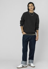 Pull en maille gris foncé texturé, coupe oversize, associé à un jean bleu délavé ample avec revers pliés, et des baskets blanches avec des accents noirs.