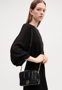 Geantă de umăr din piele neagră cu textură matlasată, accesorii aurii și curea cu lanț. Formă compactă, rectangulară, cu detalii ale logo-ului pe față.