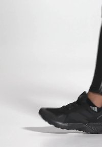 Baskets de sport noires avec une tige texturée, lacets, et une semelle épaisse, associées à des leggings noirs sur un fond clair.