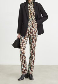 Zwarte blazer over een multikleurig geometrisch patroon shirt en broek, met tinten bruin, blauw en crème. Zwarte enkelhoge laarzen met hak.