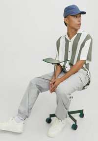 Grøn- og hvidstribet kortærmet skjorte, grå bukser, hvide sneakers, blå kasket; siddende på en grøn stol med en glat overflade.