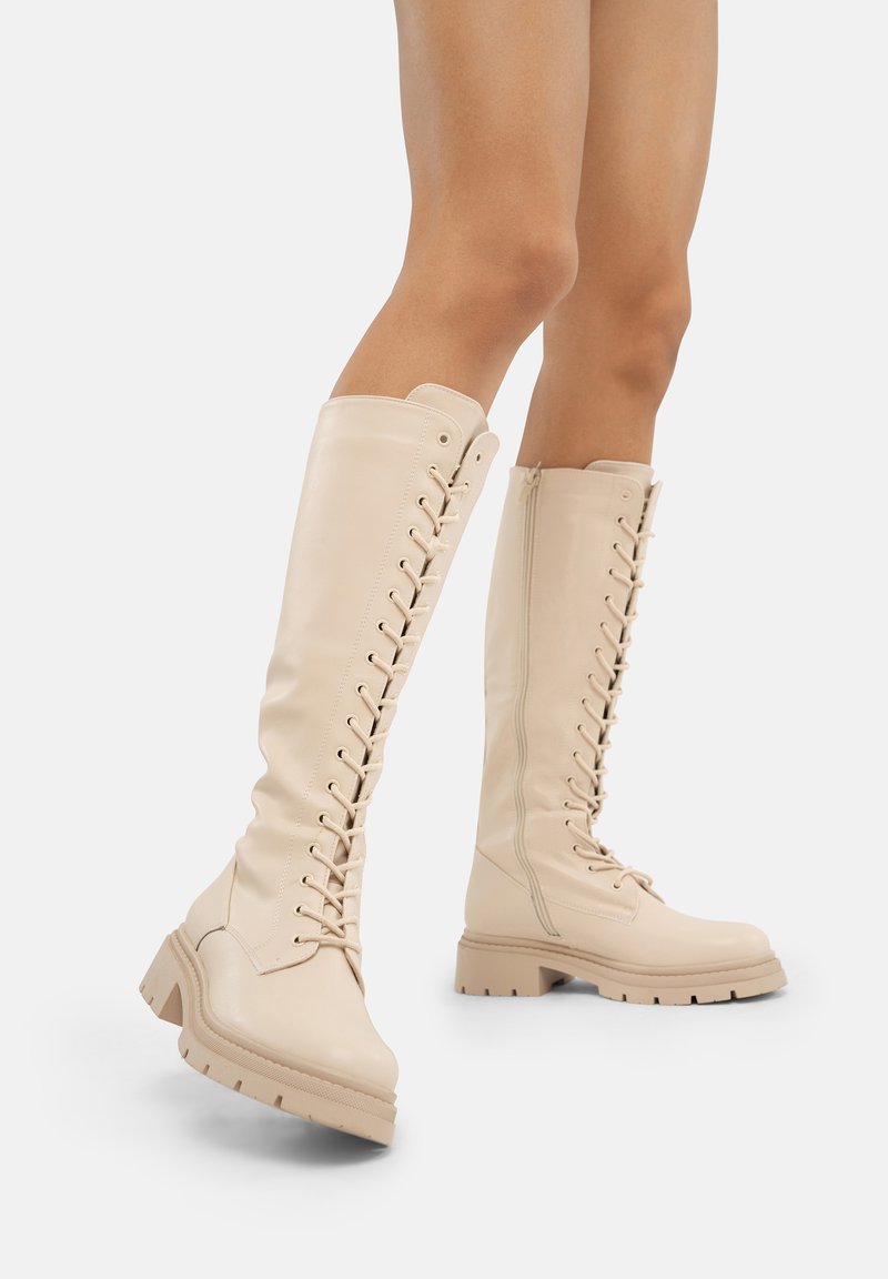 Botas de cuero beige hasta la rodilla con suela gruesa, presentan un diseño de cordones en la parte frontal y cremallera lateral para un fácil uso. Textura suave.