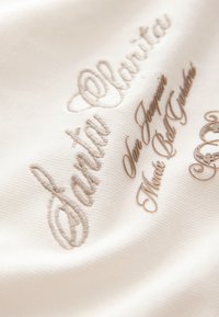 Ricamo su tessuto crema con il testo "Santa Clarita" e "San Joaquín, Moos Bel Garden" in caratteri corsivi e serif color marrone chiaro.