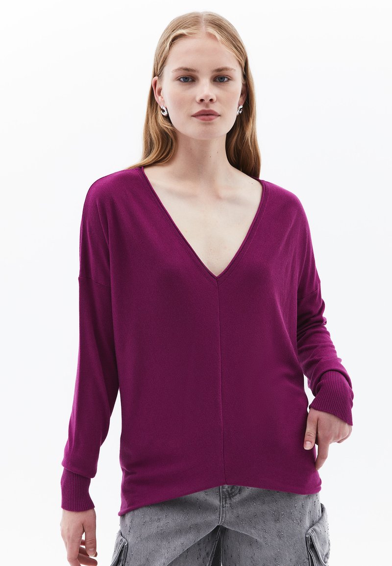 Maglione a maniche lunghe a scollo a V di un vivace magenta. Materiale morbido con polsini a coste, vestibilità ampia e orlo leggermente curvo. Abbinato a pantaloni cargo grigi.