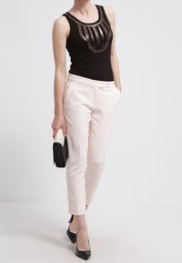 Zwarte tanktop met een pailletten detail, gecombineerd met op maat gemaakte lichtroze broeken. Model houdt een zwarte clutch vast en draagt zwarte hakken.