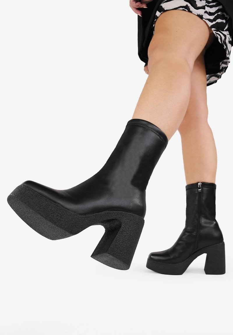 Bottines noires en cuir lisse, avec une semelle de plateau texturée et un talon bloc épais. Détail de fermeture éclair sur le côté.