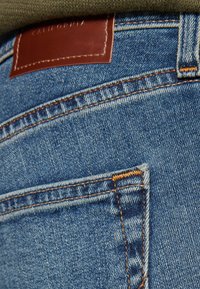 Modri denim kavbojke z rumenim šivom, ki imajo rjav usnjen značko z napisom "CALIFORNIA" in posebne podrobnosti na zadnjem žepu.