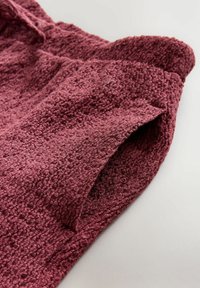 Strukturierter Stoff in tiefburgunder Farbe, der eine weiche, plüschige Oberfläche mit einem gerollten Saum und einem flachen Taschen-Detail an der Seite präsentiert.