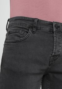 Jeans de mezclilla negra con un diseño clásico de cinco bolsillos, cierre de botón metálico y un sutil detalle de costura. Textura suave, corte regular.