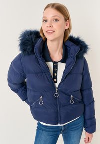 Giacca a vento blu navy con cappuccio in pelliccia sintetica, tasche con zip e dettagli in metallo argento. Presenta una silhouette aderente e un design trapuntato.