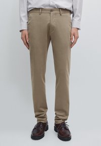 Pantaloni chino beige in cotone con vestibilità slim, con chiusura a zip, bottone e tasche laterali, abbinati a scarpe in pelle marrone scuro.
