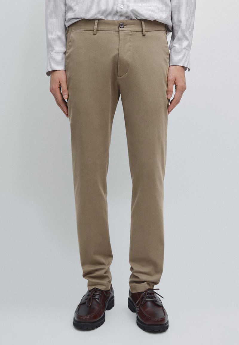 Pantaloni chino beige in cotone con vestibilità slim, con chiusura a zip, bottone e tasche laterali, abbinati a scarpe in pelle marrone scuro.