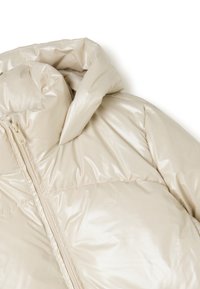 Chaqueta de plumas beige con un acabado brillante, que presenta un cierre de cremallera y un gran cuello acolchado. Textura suave y acolchada con costuras visibles.