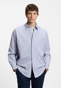 Młody mężczyzna o kręconych włosach, ubrany w jasnoniebieską koszulę zapinaną na guziki, na białej podkoszulce i ciemnych spodniach, stojący z rękami w kieszeniach.