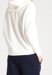 Witte capuchontrui met een zachte textuur, voorzien van een kangoeroezak en geribbelde manchetten, gecombineerd met donkerblauwe broek.