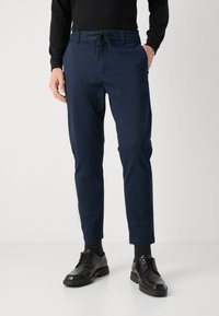 Navy blue tapered pants with an elastic waistband and drawstring, featuring side pockets and a smooth texture, paired with black dress shoes.