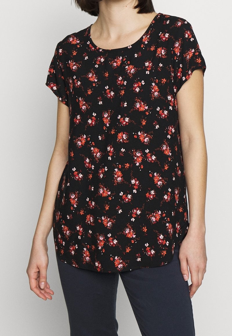 Femme portant un chemisier noir à manches courtes avec un motif floral rouge et blanc, assorti à un pantalon foncé sur un fond uni.