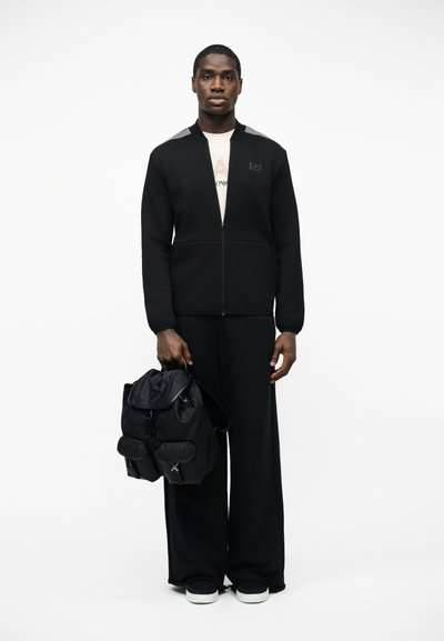 Άνδρας που φοράει μαύρο μπουφάν με φερμουάρ, φαρδιά μαύρη παντελόνια, λευκά αθλητικά παπούτσια, κρατώντας ένα μαύρο σακίδιο πλάτης μπροστά σε απλό λευκό φόντο.