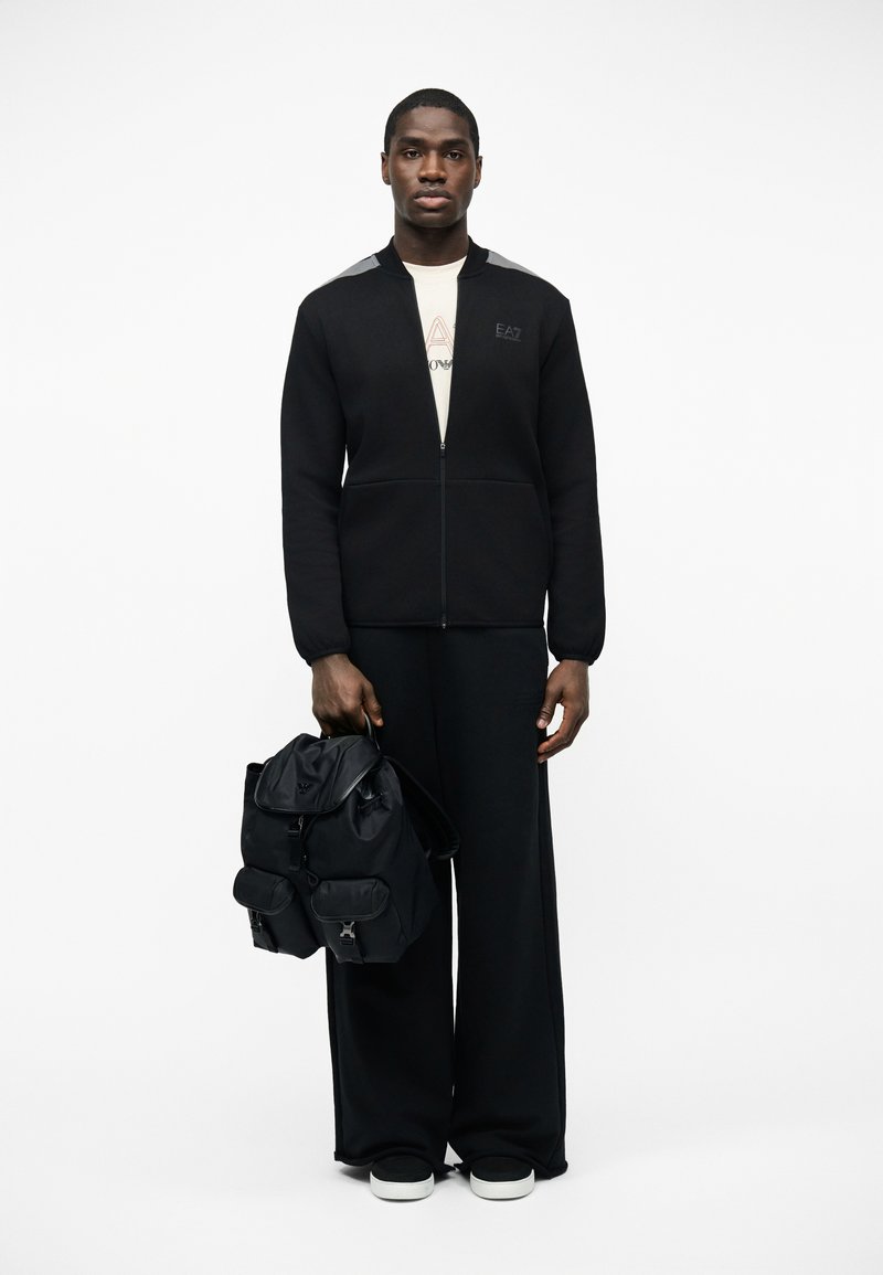 Uomo che indossa una giacca nera con cerniera, pantaloni neri larghi, sneakers bianche, che tiene uno zaino nero su uno sfondo bianco uniforme.