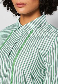 Camicia bianca con strisce verticali verdi; presenta un colletto a punta, chiusura a bottoni e dettagli testurizzati verdi lungo la patta.
