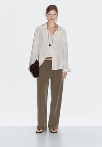 Light beige button-up shirt, loose-fitting olive green pants, dark brown clutch, and a long black pendant necklace. Flat sandals visible.