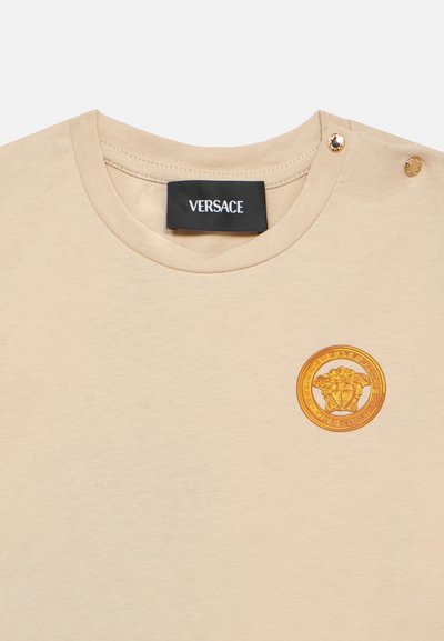 Μπεζ βαμβακερό t-shirt με στρογγυλή λαιμόκοψη, χρυσά διακοσμητικά κουμπιά στον ώμο και χ ρυσό έμβλημα Medusa στο στήθος.