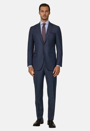Homme debout face à l'appareil portant un costume bleu foncé à deux boutons, une chemise bleu clair, une cravate à motifs et des chaussures habillées noires sur un fond uni.