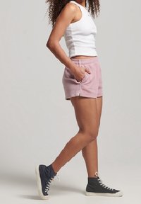 Shorts de algodón rosa con bolsillos laterales, combinados con una camiseta sin mangas blanca ajustada y zapatillas negras de caña alta. La modelo está de perfil sobre un fondo neutro.