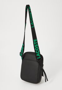Borsa a tracolla nera con superficie texturizzata, chiusura con zip e tracolla con logo verde. Forma quadrata con tasca frontale. Tracolla regolabile.