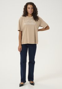 T-shirt oversize beige en coton à manches courtes avec du texte blanc, associé à un jean droit bleu marine et des chaussures noires à talons.